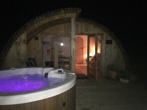 A wooden cabin with a hot tub in the foreground and a cozy interior visible through the open door.