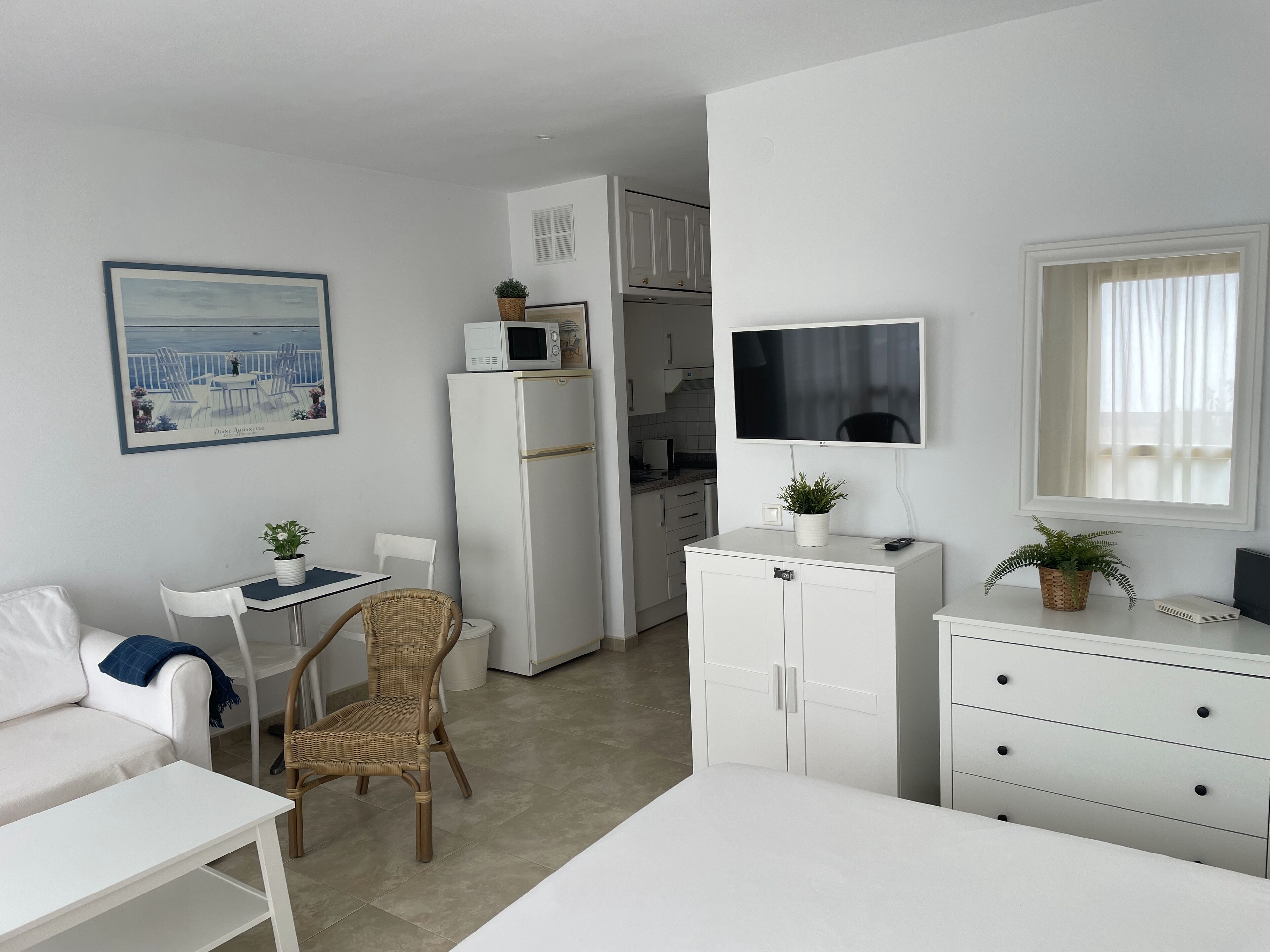 Quarto moderno e minimalista com sofá branco, mesa de jantar, cadeira de vime, cozinha compacta, televisão e uma janela grande.
