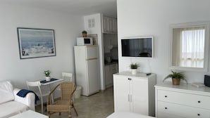 Una camera moderna e minimalista con un divano bianco, un tavolo da pranzo, una sedia in vimini, un angolo cottura, una televisione e un'ampia finestra.