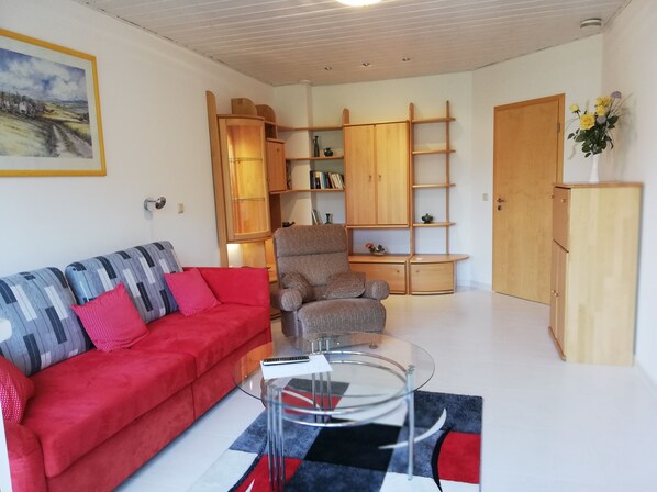 Un salon avec un canapé rouge, un fauteuil gris, une table basse en verre, des étagères en bois et une porte.