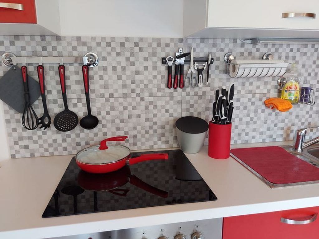 A kitchen with a red pot on a black induction cooktop, a hanging rack with utensils, and a red cutting board on the counter.