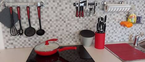 A kitchen with a red pot on a black induction cooktop, a hanging rack with utensils, and a red cutting board on the counter.