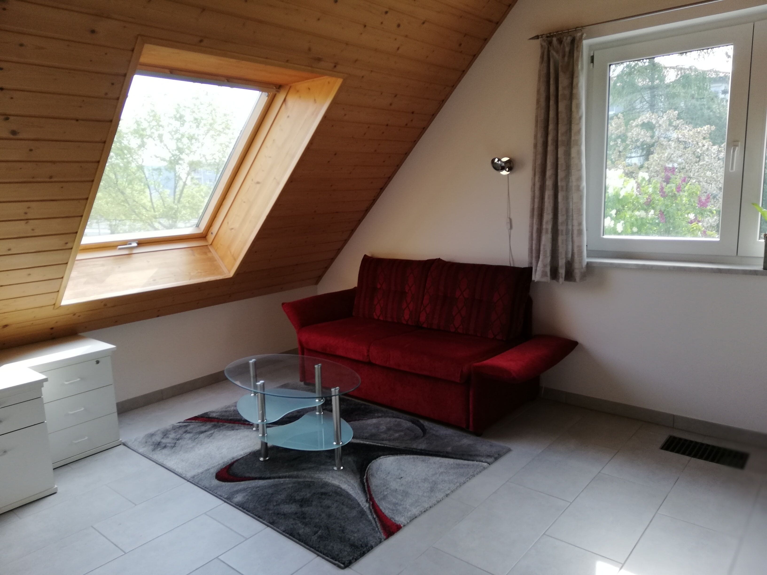 A room with a sloped ceiling, a red sofa, a glass coffee table, and a white cabinet.