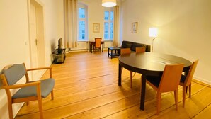 A spacious room with a wooden floor, a round dining table, and several chairs. There is a sofa and a small table with a lamp. The room has large windows with curtains. - BerlinLUX Apartments - City (Berlin)
