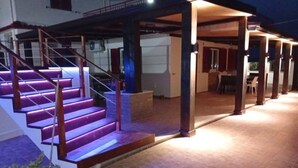 Ein überdachter Außenbereich mit einer Treppe, die zu einer erhöhten Ebene führt, einem Essbereich und Blick auf ein Gebäude im Hintergrund.