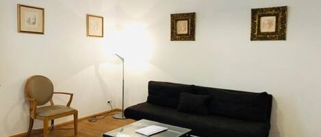 Uma sala de estar com um sofá preto, uma cadeira de madeira, uma mesa de centro de vidro e quadros nas paredes.