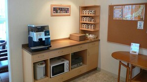 Una kitchenette con cafetera, microondas y cajones para guardar cosas. Hay un tablón de anuncios con mapas y avisos, y una mesita con un cartel.