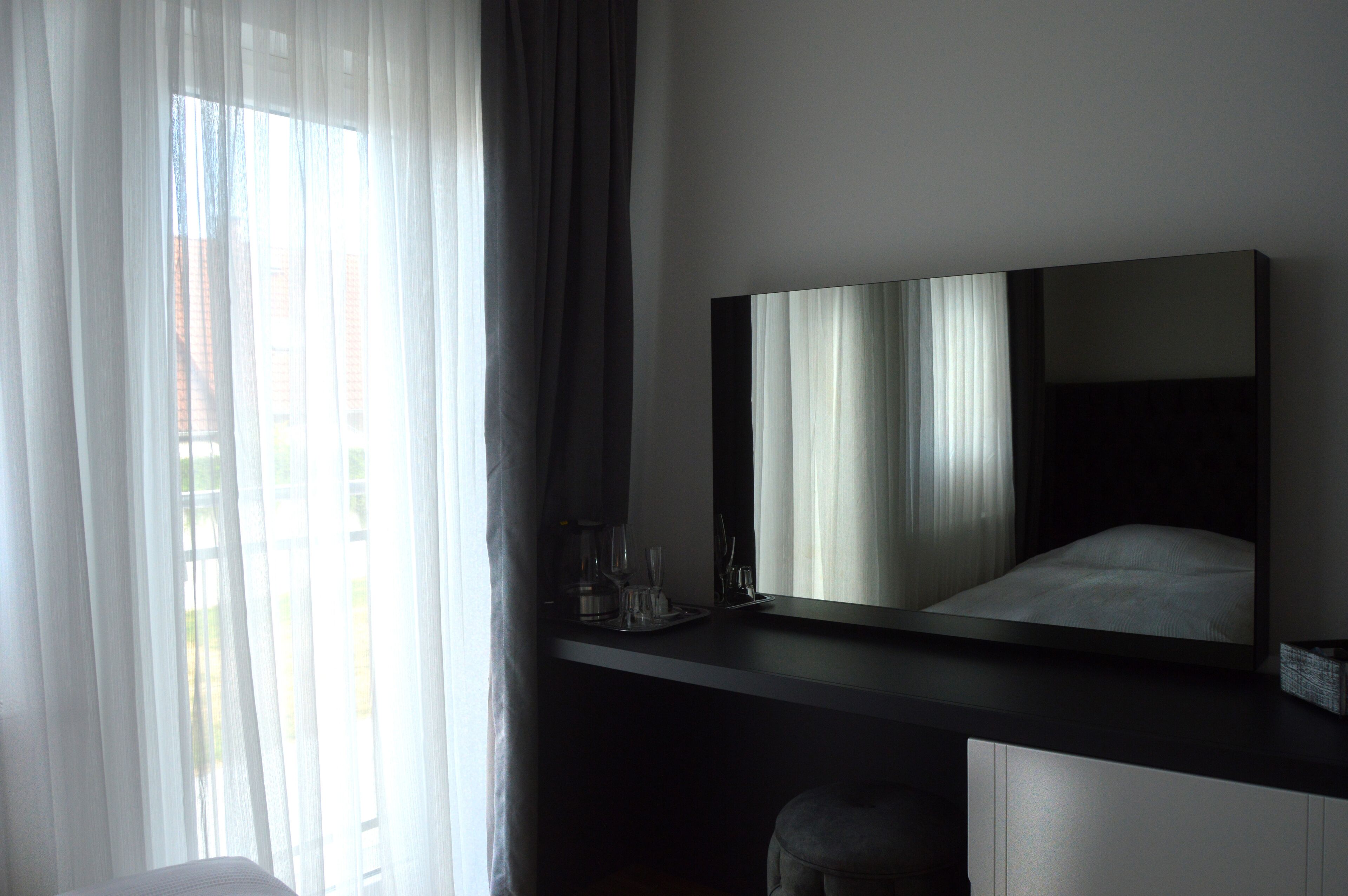 Una habitación de hotel moderna con una cama grande, una mesita de noche con lámpara y un ventanal con cortinas translúcidas.