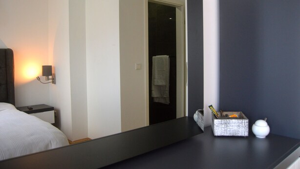 A hotel room with a bed, bedside table, nightstand, mirror, and a small table with a box and a white object.