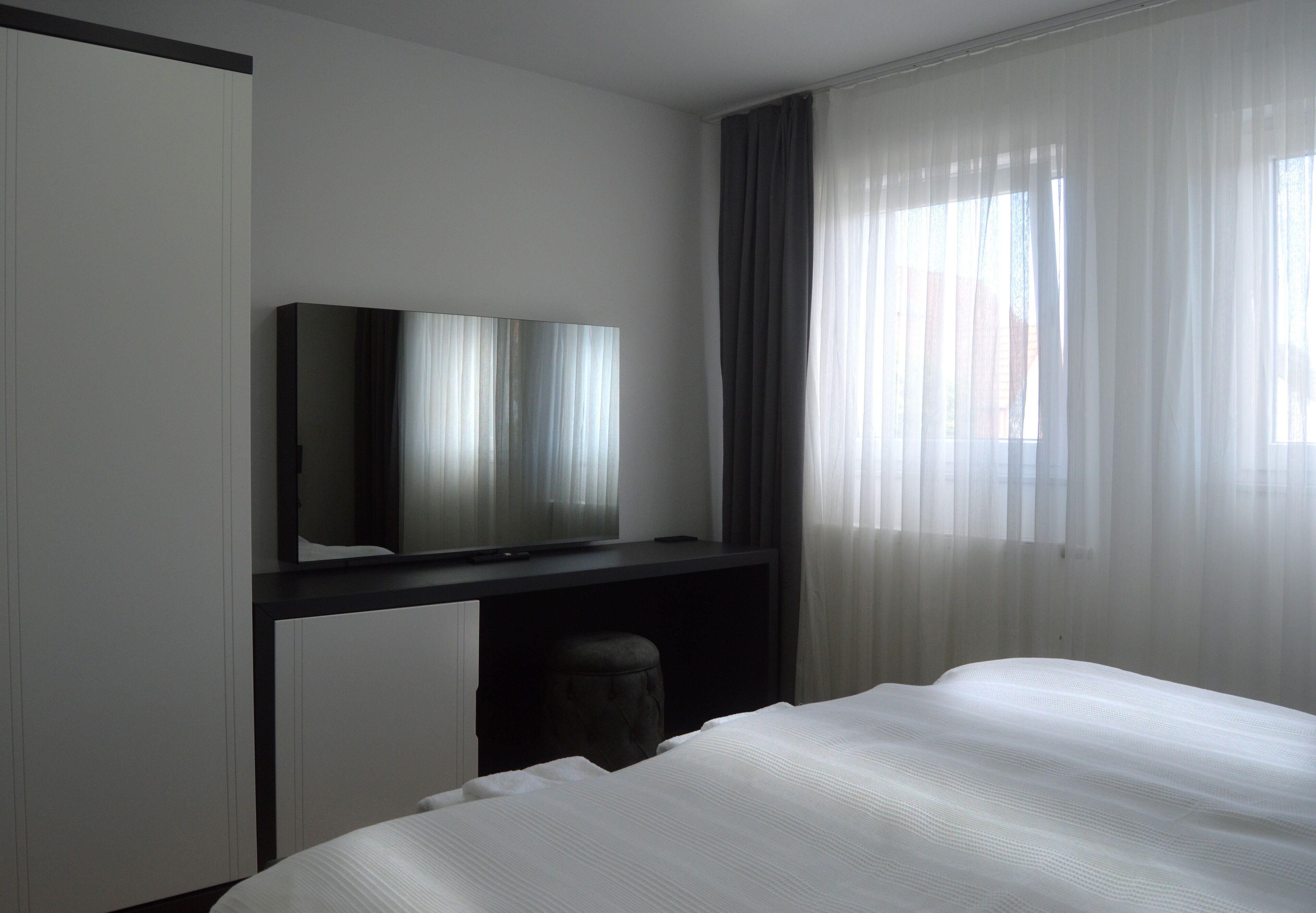 A hotel room with a bed, a TV on a stand, a desk, and a window with sheer curtains.