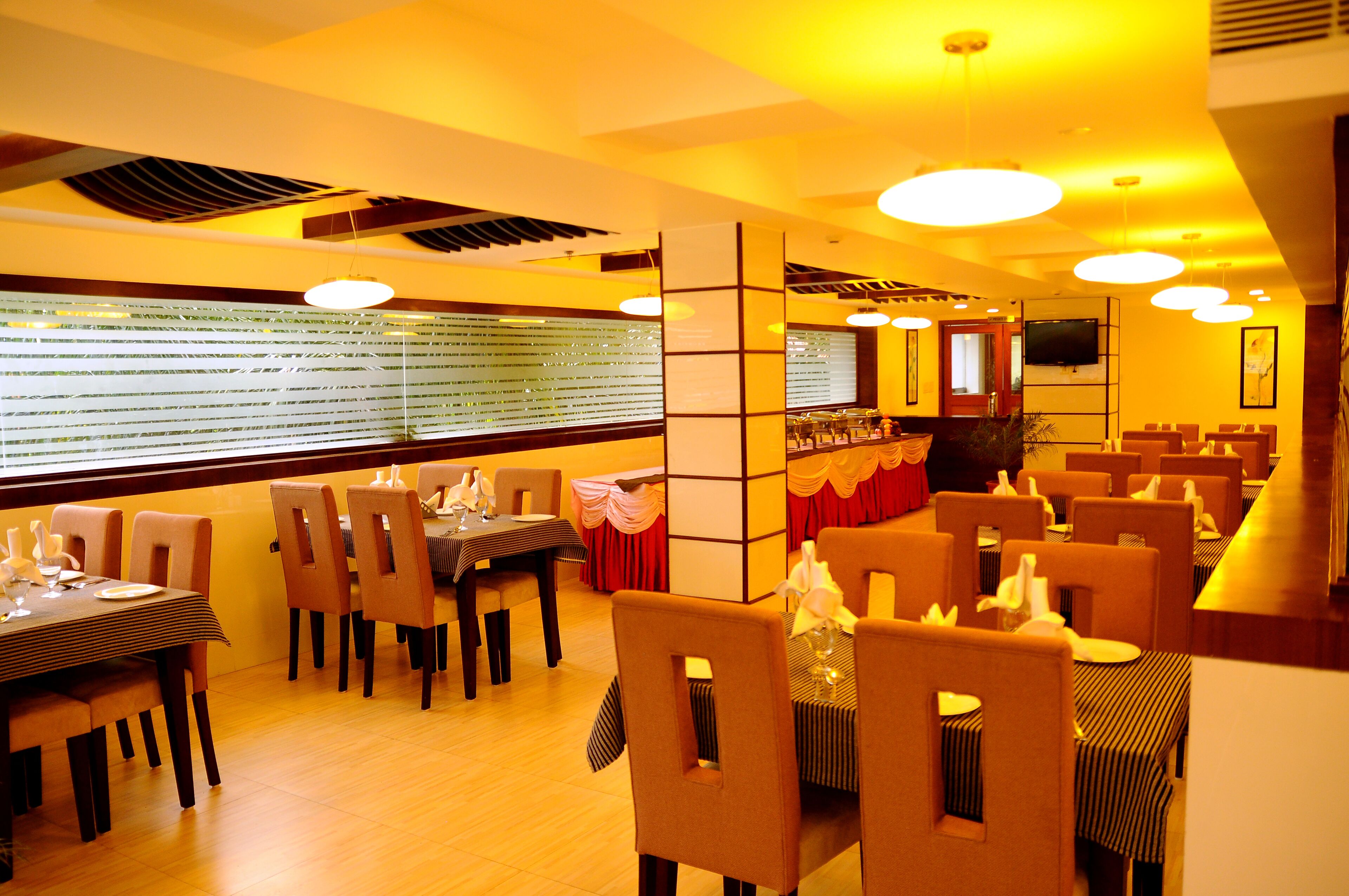 A restaurant with multiple dining tables and chairs, a television mounted on the wall, and a window with blinds.