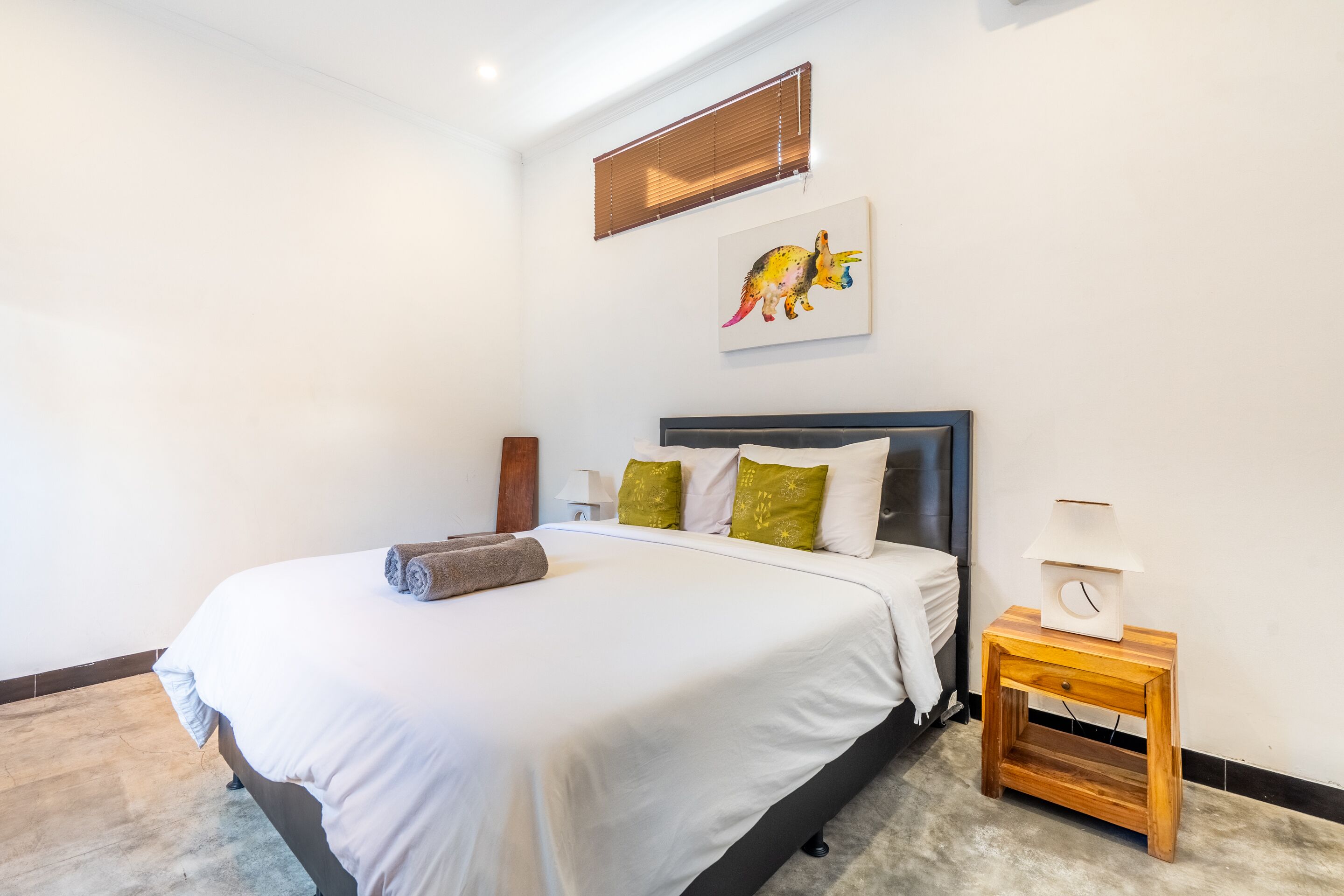 A neatly made bed with white linens and a brown towel, a bedside table with a lamp, and a wall-mounted artwork of a dinosaur.