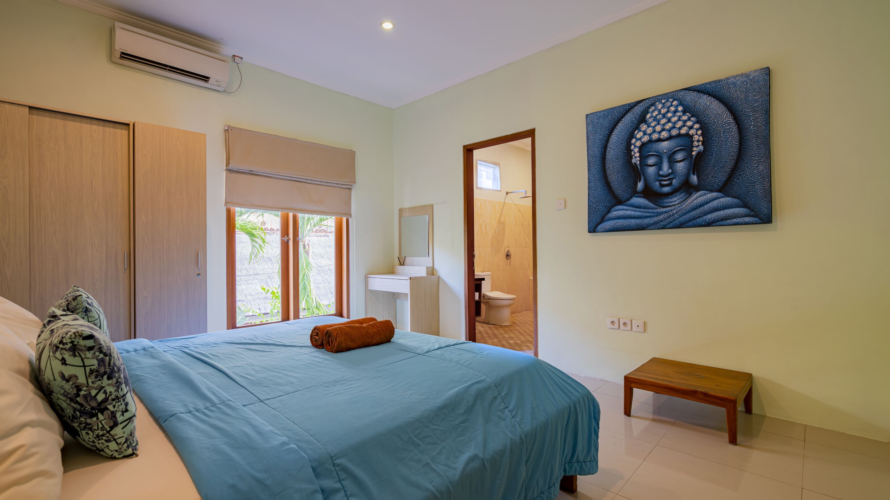 A bedroom with a bed, a bench, a bathroom with a toilet and sink, and a large Buddha painting on the wall.
