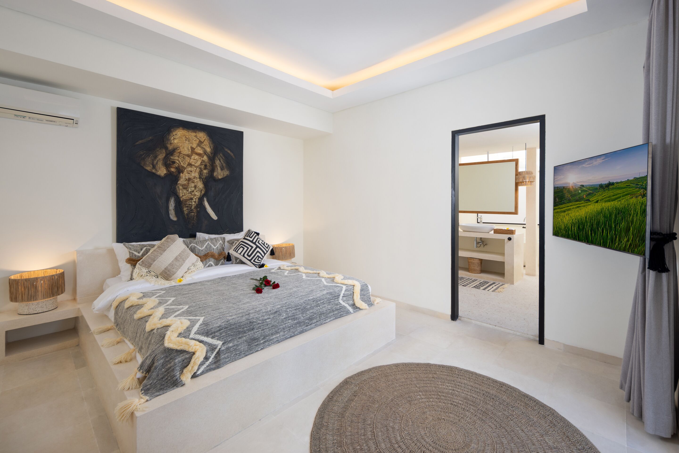 Ein modernes Schlafzimmer mit einem großen Bett, einem Gemälde, einem Fernseher und einem durch eine offene Tür einsehbaren Badezimmer.