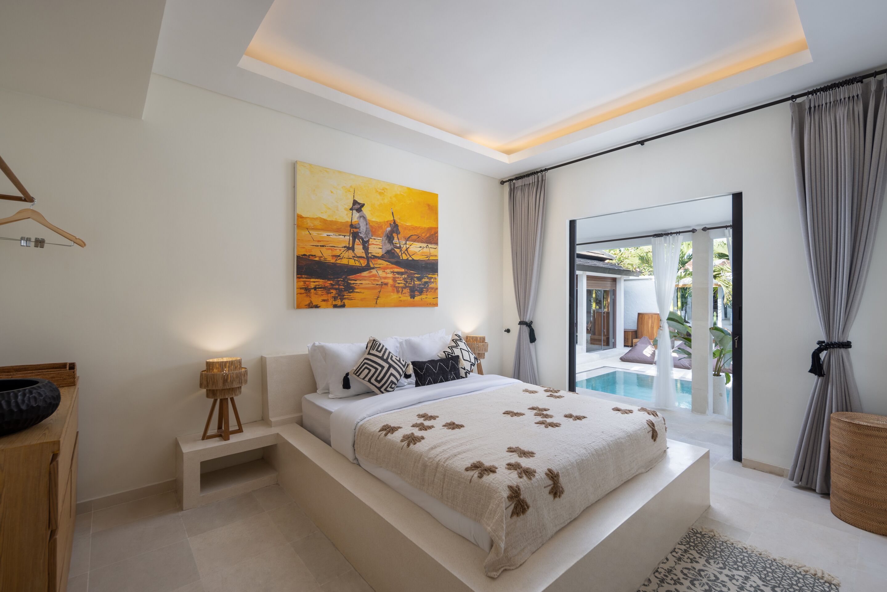 A modern bedroom with a large bed, a painting, a view of a pool, and a wooden side table.