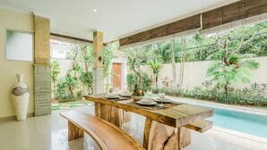 Un espace repas avec une table en bois dressée pour quatre personnes, entouré d’une végétation luxuriante et d’une piscine.