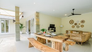 Un grand espace dédié à la salle à manger, avec une table et des bancs en bois, un canapé en osier et un téléviseur en arrière-plan.