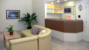 Un área de recepción con un sofá color beige, una planta en maceta y un mostrador de recepción con un reloj y un cuadro enmarcado encima.