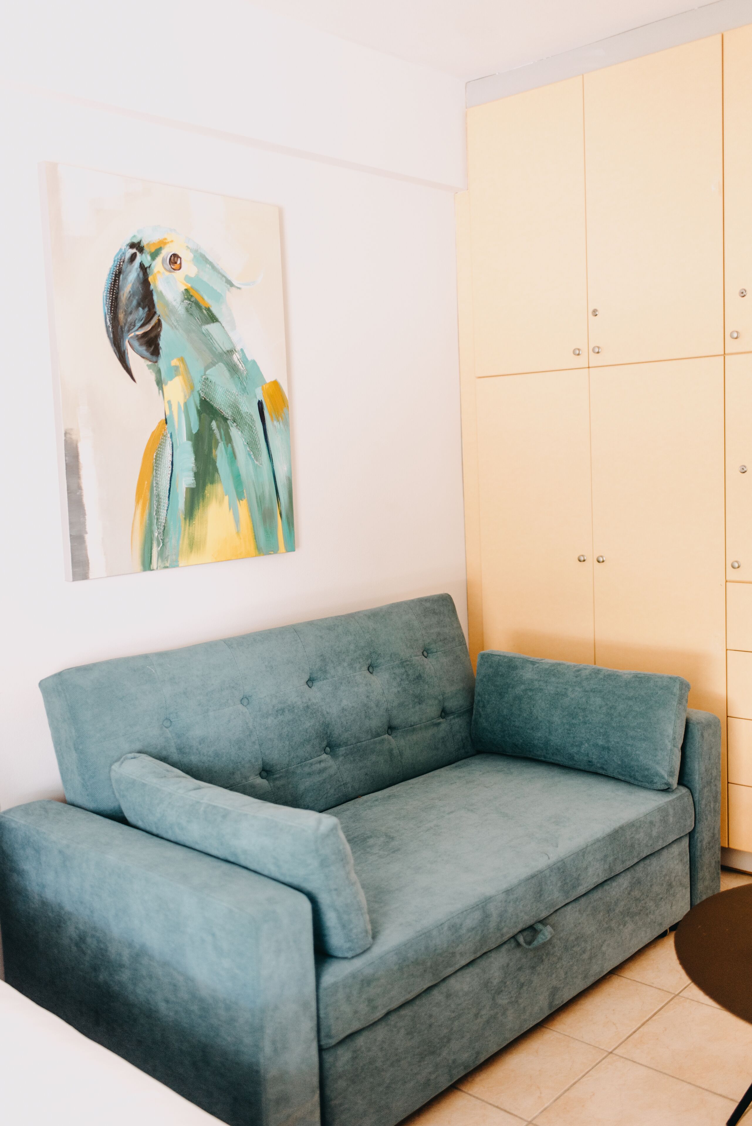 Un canapé de couleur turquoise, une armoire de couleur claire et un tableau représentant un perroquet accroché au mur.