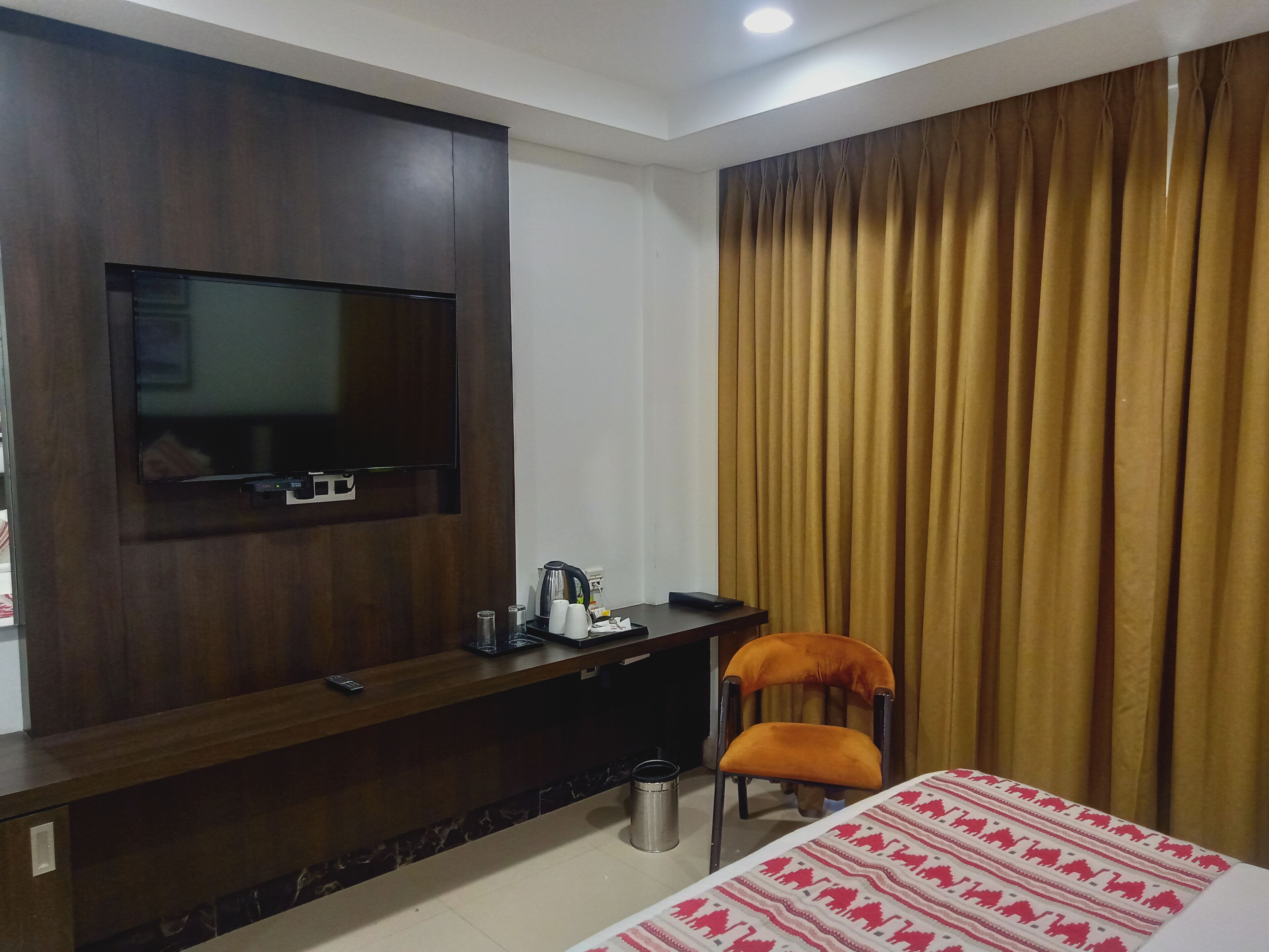 A hotel room with a dark wood TV stand, a bed with a patterned bedspread, an orange chair, and a large window with curtains.