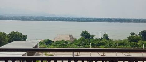 A view of a river with a distant shoreline, a pool area with a geometric fence, and greenery in the foreground.