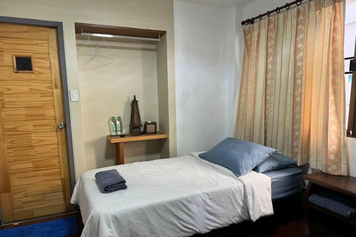 A small, neatly arranged bedroom with a bed, a wooden wardrobe, a small table with a lamp, and a curtain.