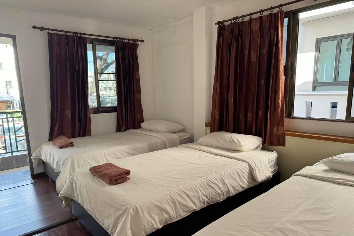 A hotel room with three single beds, each with a white mattress and pillows. There are brown curtains on the windows, and the room has a wooden floor.