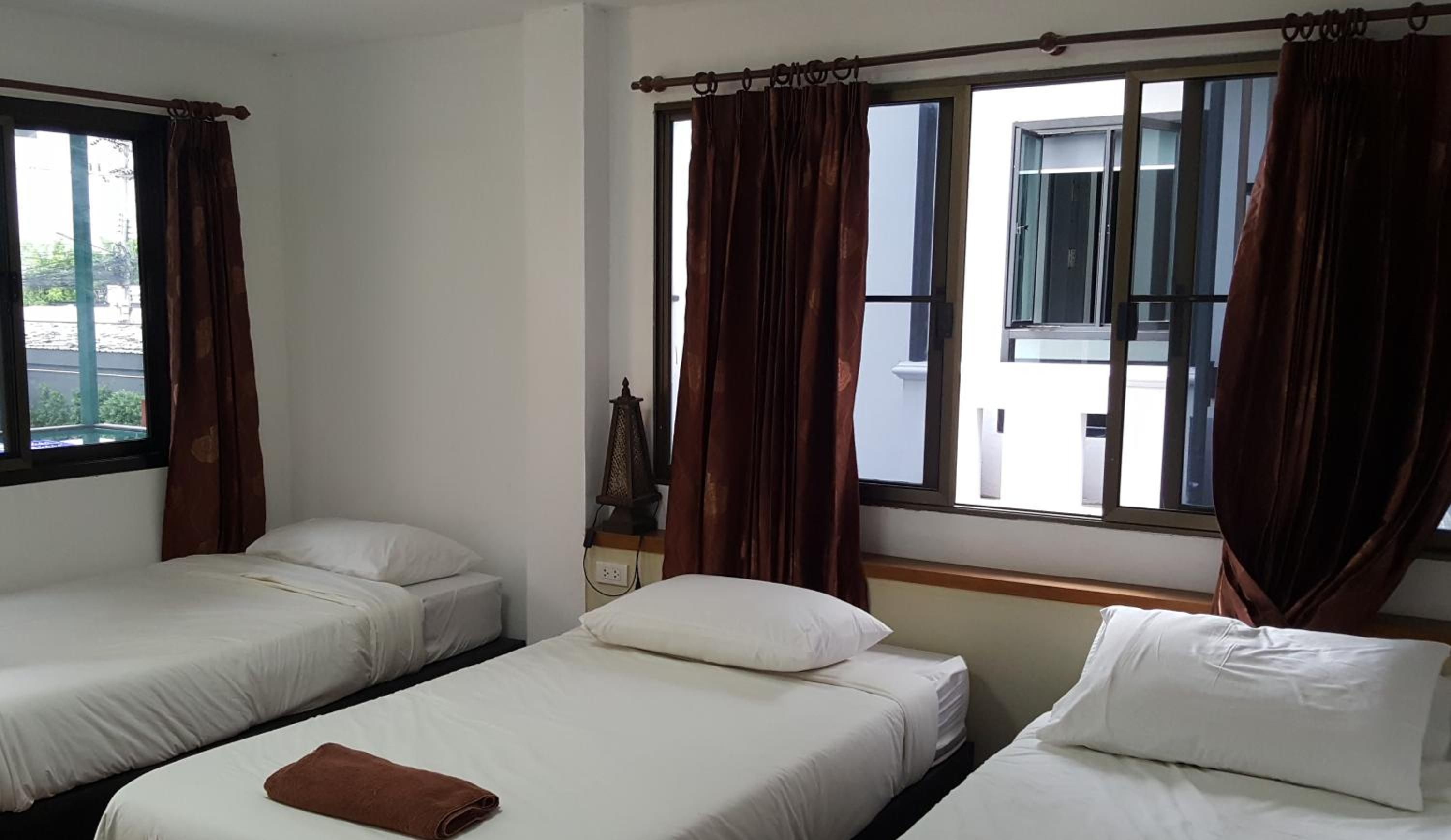 A room with three single beds, a window with curtains, and a small table with a lamp.