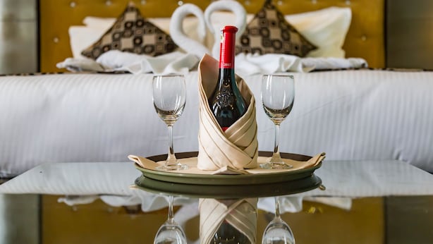 A bed with a tufted headboard, two pillows with a geometric pattern, and a bottle of wine with two glasses on a tray.