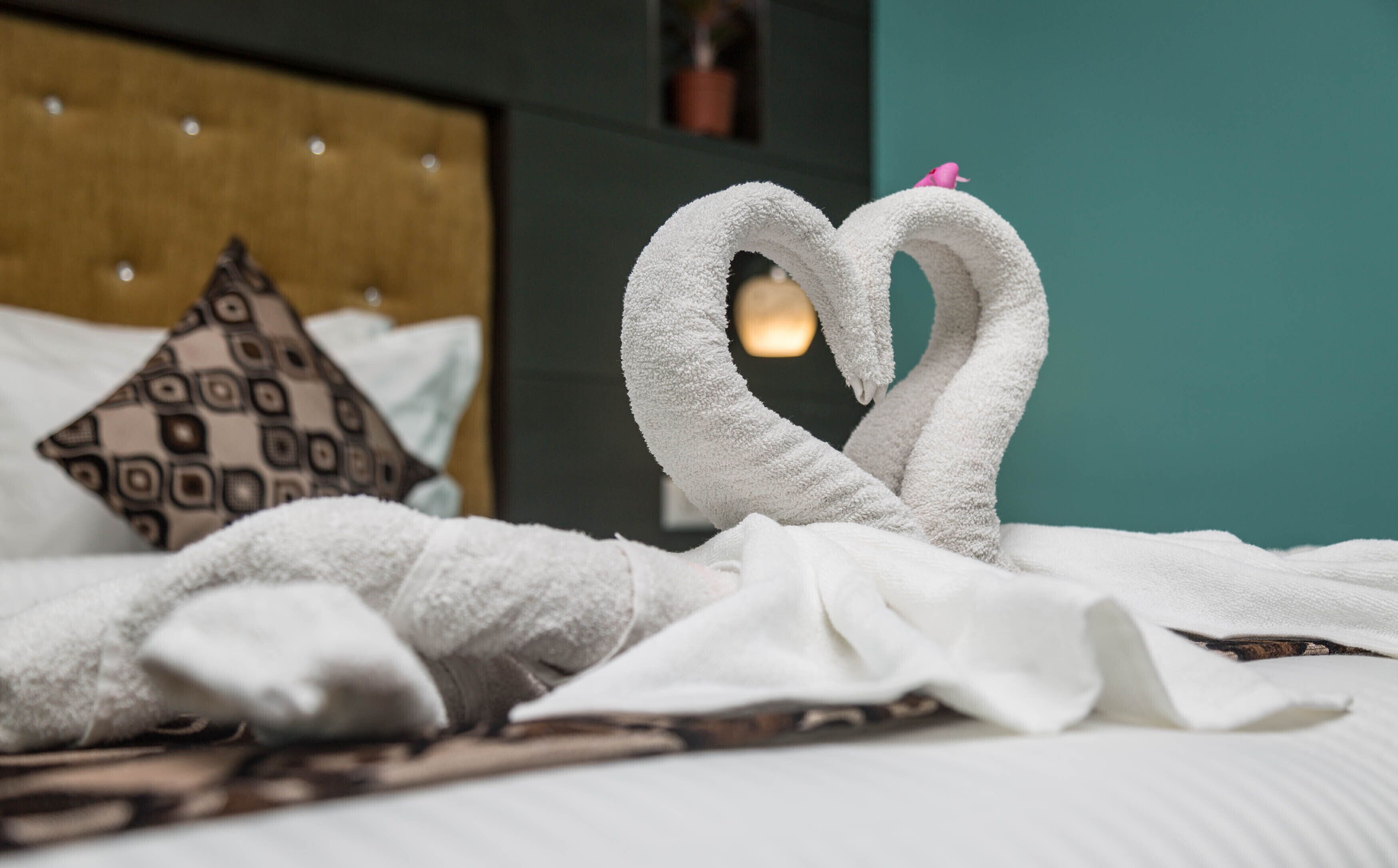 A bed with white towels arranged in a heart shape, a patterned pillow, and a teal wall in the background.