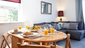 A dining area with a round wooden table, plates with croissants and fruit, and glasses of orange juice. There is a grey sofa and a mirror on the wall. - Great Havers (Bishop's Stortford)