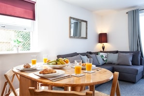 A dining area with a round wooden table, plates with croissants and fruit, and glasses of orange juice. There is a grey sofa and a mirror on the wall. - Great Havers (Bishop's Stortford)
