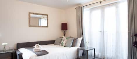A hotel room with a large bed, a bedside table, a nightstand, a mirror, a chandelier, and a view of the outdoors through large windows.