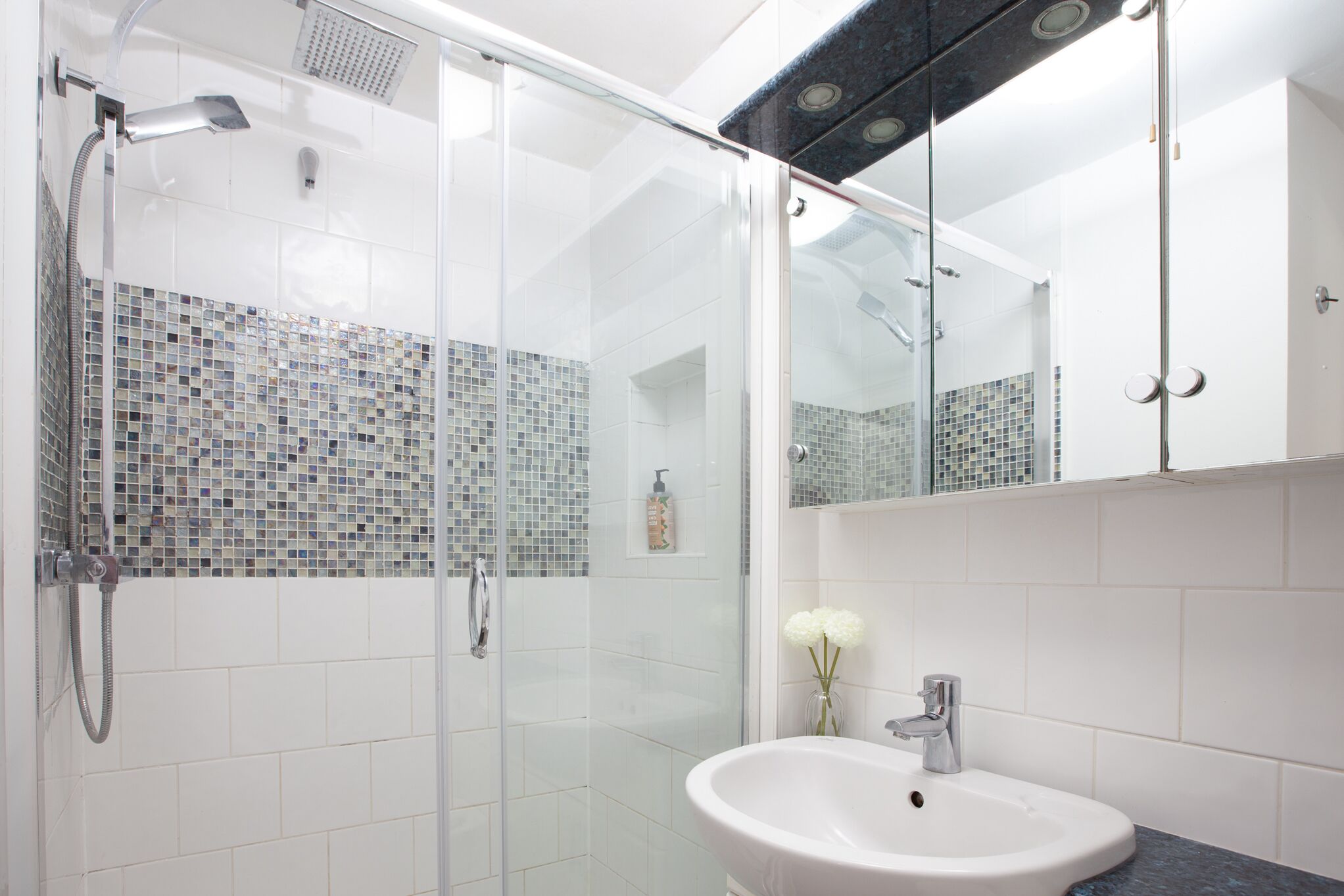 Une salle de bain moderne avec une douche à l’italienne en verre, un lavabo blanc et un mur recouvert de carreaux.