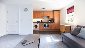 A compact living space with a kitchenette, a washing machine, a sofa, and a table with a laptop. - Great Havers (Bishop's Stortford)