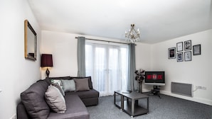 A modern living room with a grey sofa, a small table, a TV displaying 'NETFLIX', and a chandelier. - Great Havers (Bishop's Stortford)