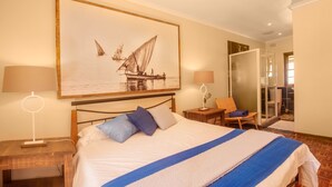 A hotel room with a bed, two bedside tables, a chair, a large painting of a sailboat, and a shower area. - Saxenburg Wine Farm (Cape Town)
