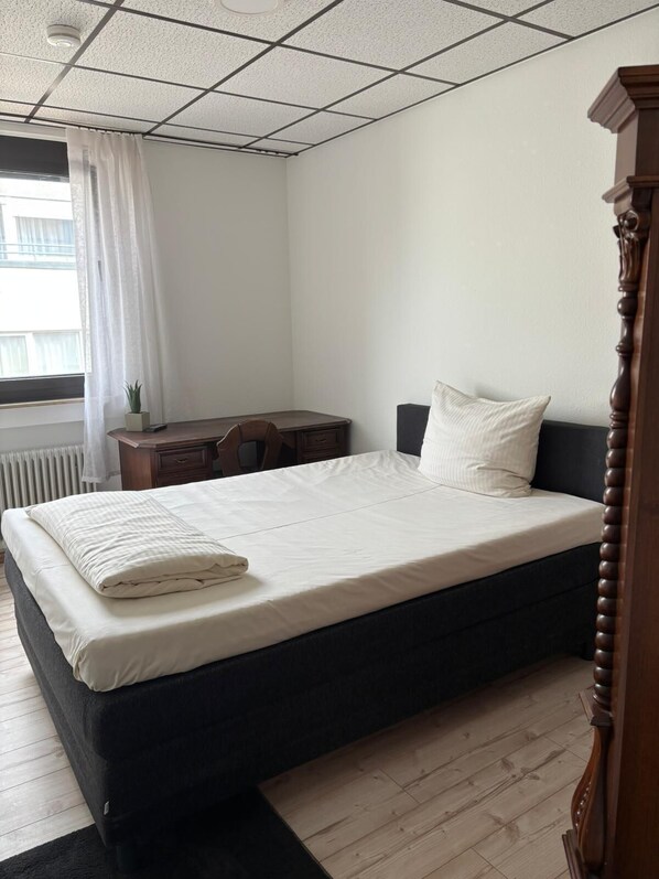 Un dormitorio con cama, escritorio, silla, radiador y ventana con cortinas.