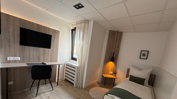 A hotel room with a bed, a desk with a chair, a TV mounted on the wall, a bedside table with a lamp, and a window with curtains. - Hotel Vi Vintage (Stuttgart)