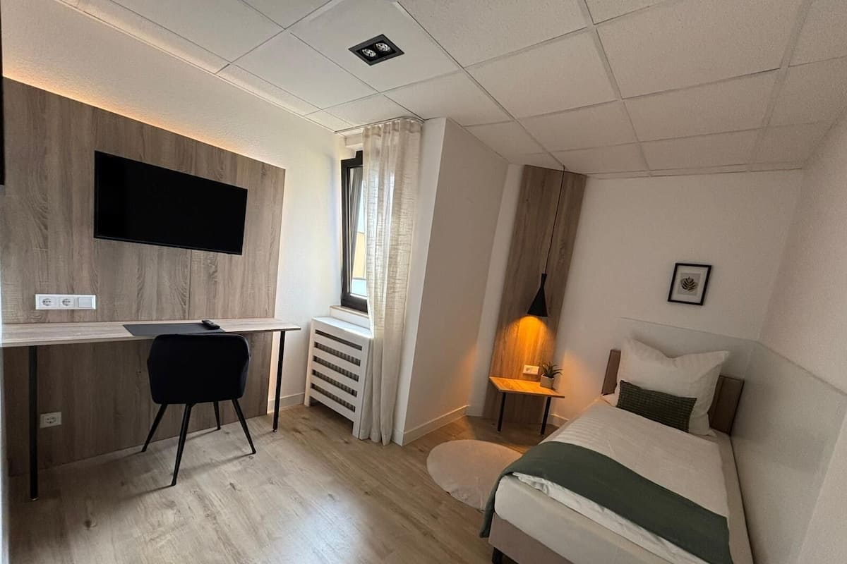 Ett hotellrum med en säng, ett skrivbord med en stol, en tv som är monterad på väggen, ett nattduksbord med en lampa och ett fönster med gardiner.