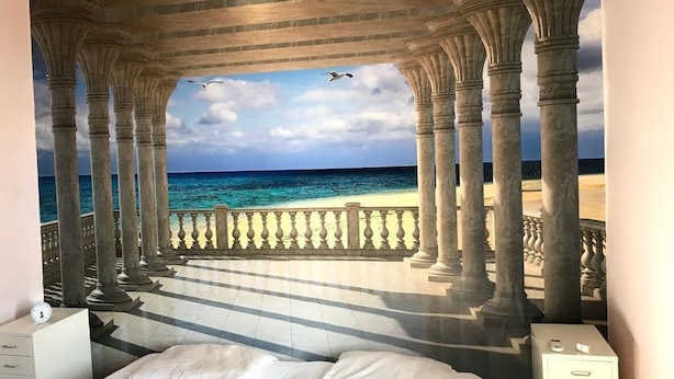A bedroom with a large bed, a nightstand, and a dresser. There is a mural of a beach scene with columns and a railing. - Villa Garibaldi. Beautiful mountain town in wine area ½ hour from Rome and beach (Cori)