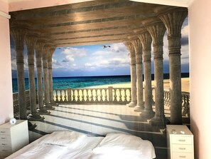 A bedroom with a large bed, a nightstand, and a dresser. There is a mural of a beach scene with columns and a railing. - Villa Garibaldi. Beautiful mountain town in wine area ½ hour from Rome and beach (Cori)