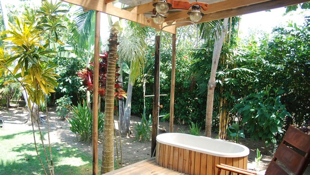 Une terrasse en bois dotée d’un jacuzzi, entourée d’une végétation tropicale luxuriante et agrémentée d’une pergola en bois ornée de guirlandes lumineuses.
