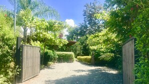 Un chemin de gravier mène à une entrée fermée par un portail, qui est elle-même entourée d’une végétation luxuriante et d’arbres.