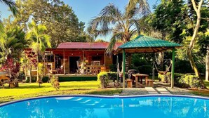 Une piscine à l’eau d’un bleu limpide, entourée d’une terrasse bordée de galets. Au fond, on aperçoit un bâtiment en bois avec un toit rouge et un espace extérieur aménagé avec un auvent.