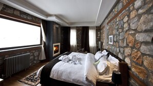 A bedroom with a stone wall, a fireplace, a large window, and a bed with white linens. - Miramonte Chalet Hotel Spa (Edessa)