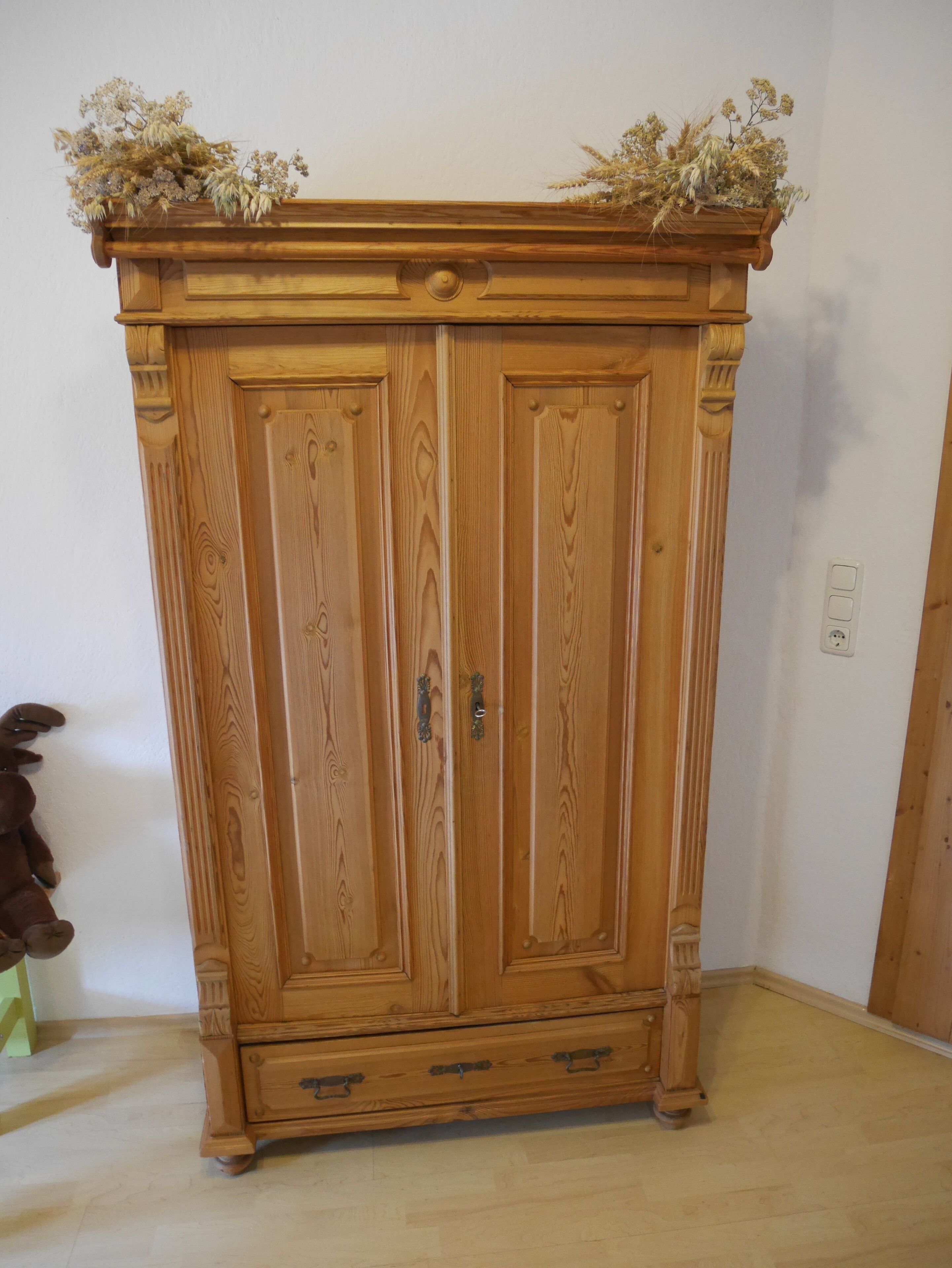 A wooden armoire with two doors and a small shelf at the top, featuring a decorative floral arrangement.