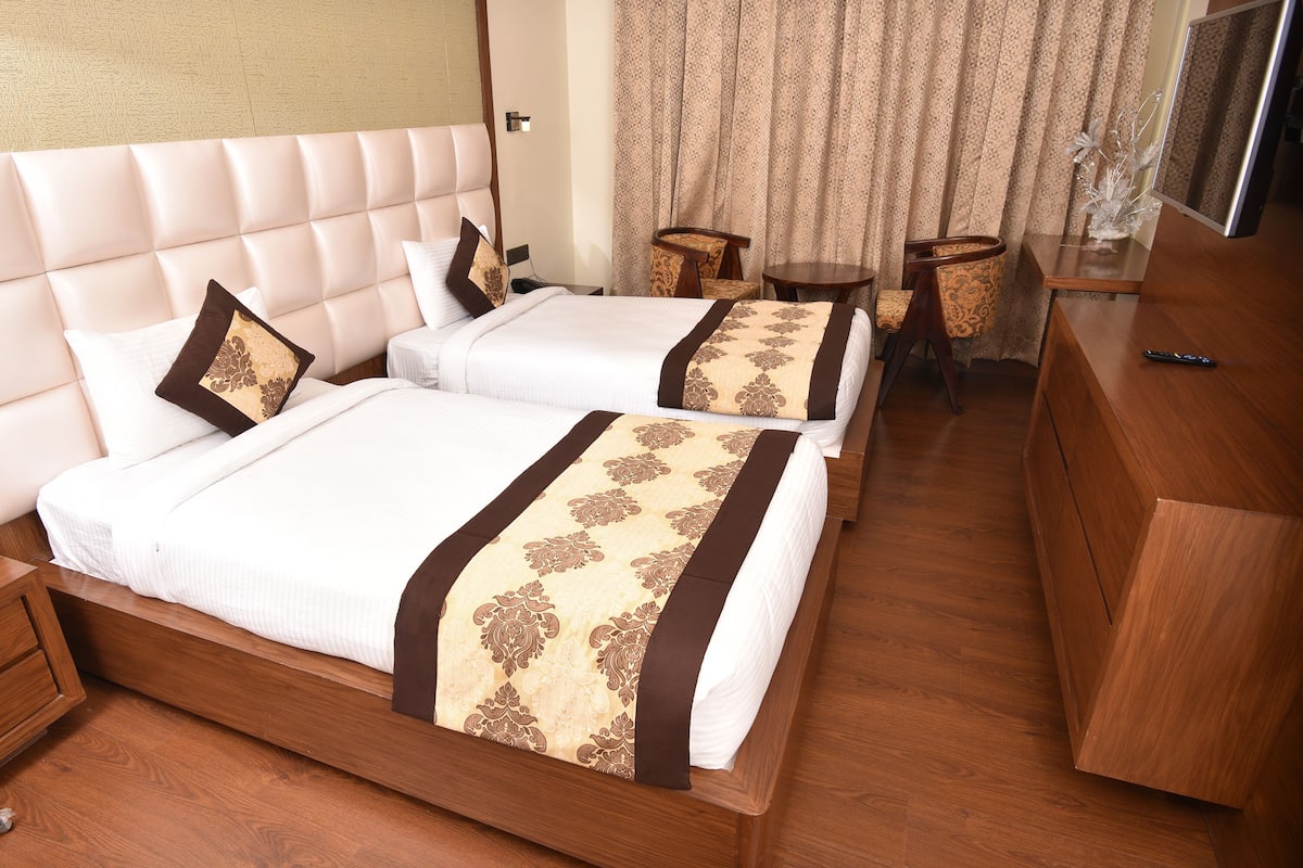 Quarto de hotel com duas camas, uma cômoda de madeira, uma mesinha e uma televisão.