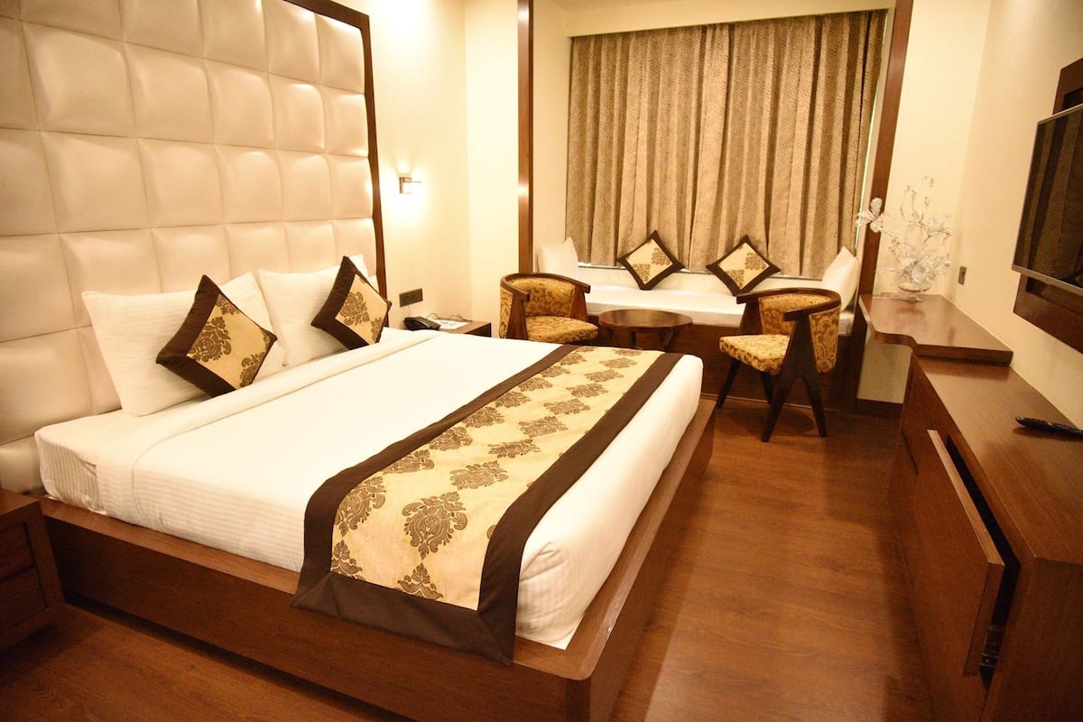 Quarto de hotel com uma cama grande, uma escrivaninha, duas cadeiras, uma televisão e uma janela com cortinas.