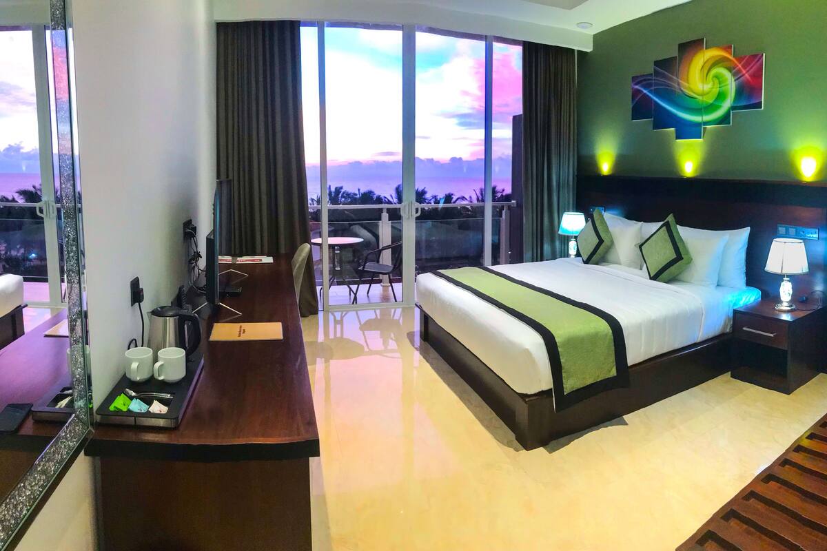 A hotel room with a large bed, a desk with a coffee maker, a chair, and a view of the cityscape through large windows.